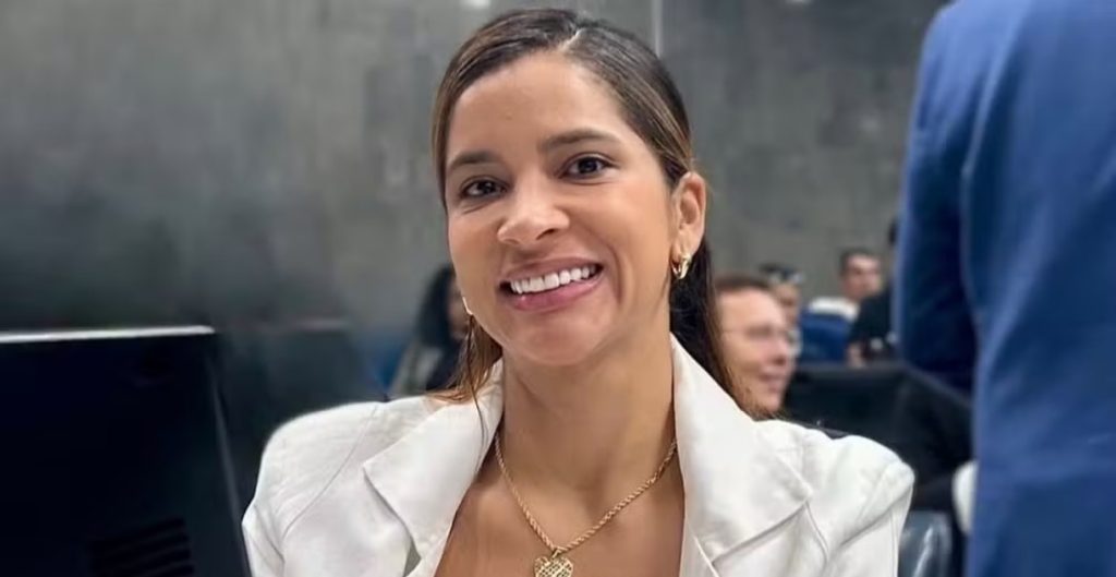 Vereadora Tatiana Medeiros sorrindo para a câmera em um ambiente interno, vestindo um blazer branco e colar dourado.