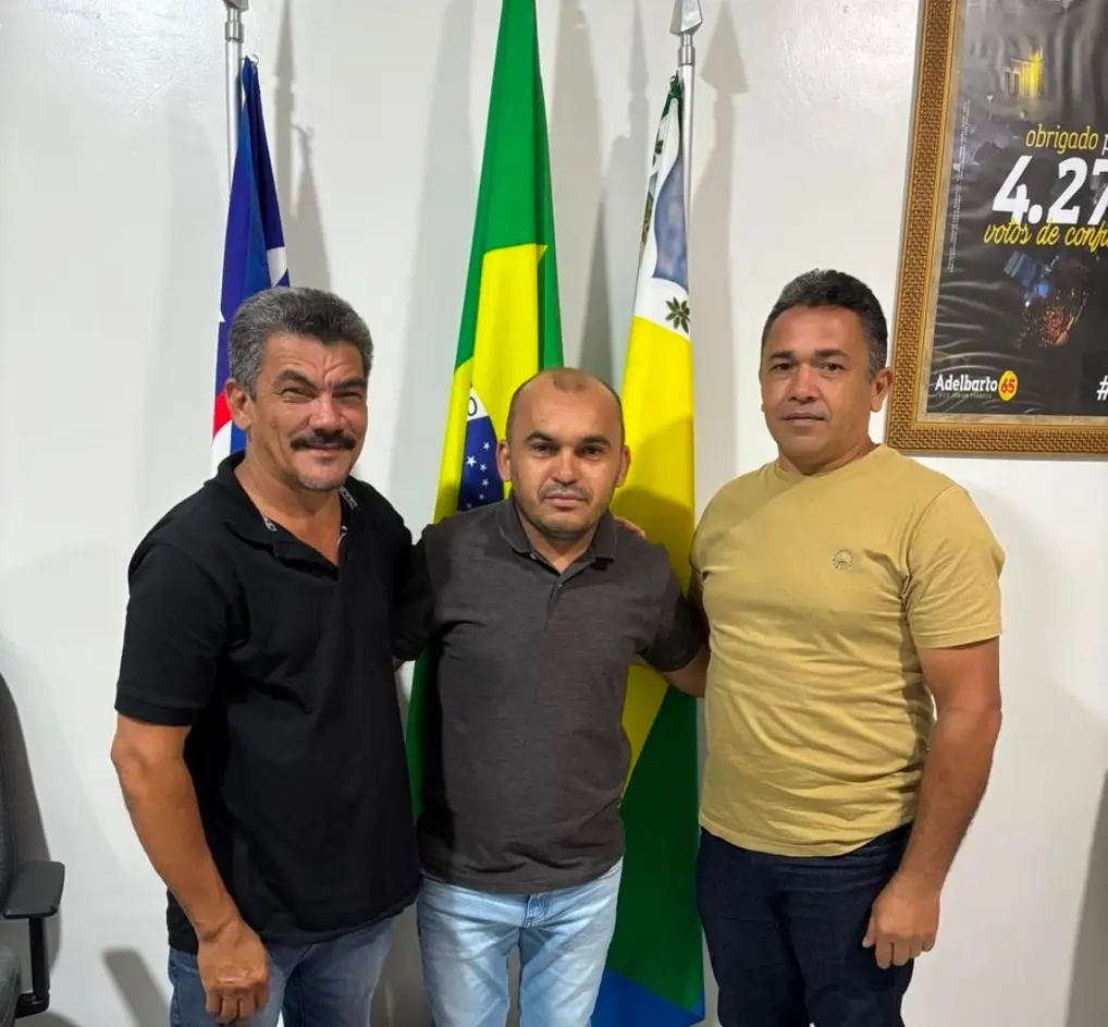 Três homens posam lado a lado e sorriem para a foto oficial. Eles estão em um ambiente interno, em pé na frente de três bandeiras (incluindo a do Brasil e a do município). A imagem registra a apresentação da nova equipe da Secretaria de Educação de São Francisco do Maranhão, com o novo secretário, professor Ozeas, e o assessor Francimilton