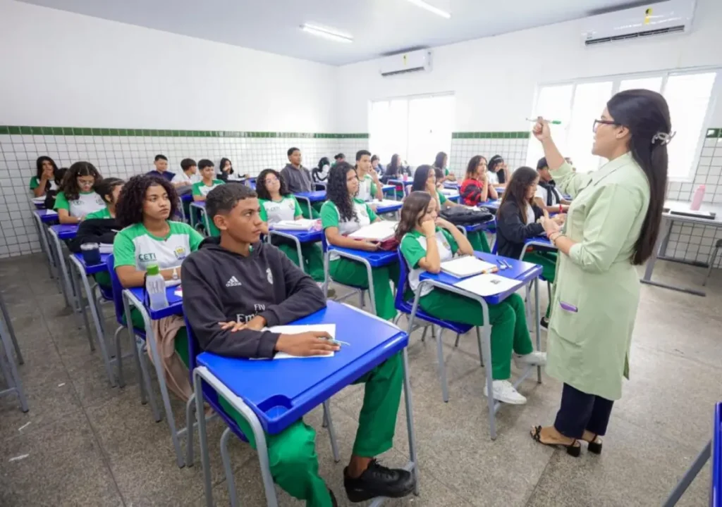 Professora ministra aula em sala da rede estadual, apontando para a lousa diante de estudantes.