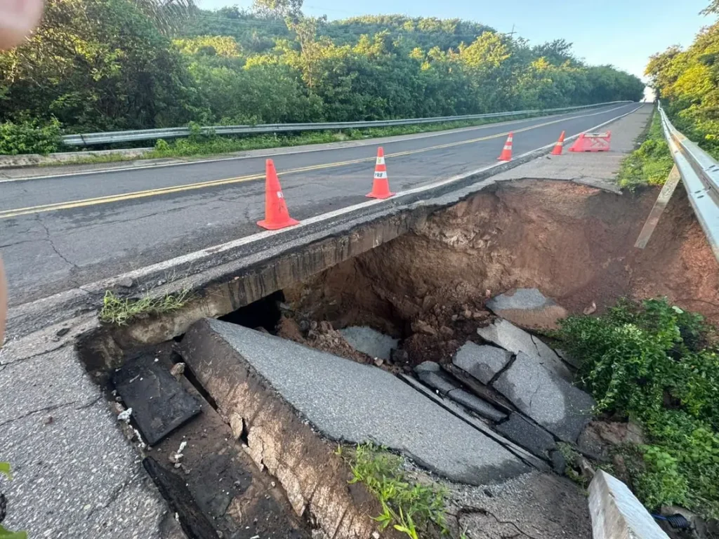 Trecho de rodovia com grande buraco aberto no acostamento direito, expondo a estrutura interna da pista e tubulação subterrânea, com quatro cones laranja sinalizando o perigo na via de tráfego