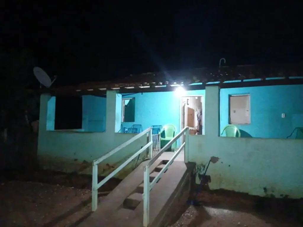 Fachada de uma casa azul com varanda e rampa de acesso à noite. A porta está aberta e uma luz ilumina a entrada.