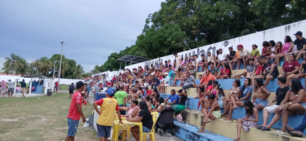 Torcedores lotam arquibancada de cimento de estádio local para assistir a um jogo de futebol sob um céu nublado.