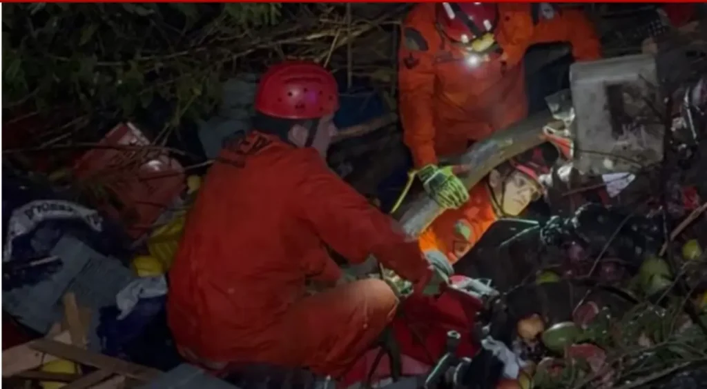 Três socorristas em uniformes laranjas e capacetes vermelhos em uma operação de resgate noturna em meio a destroços.