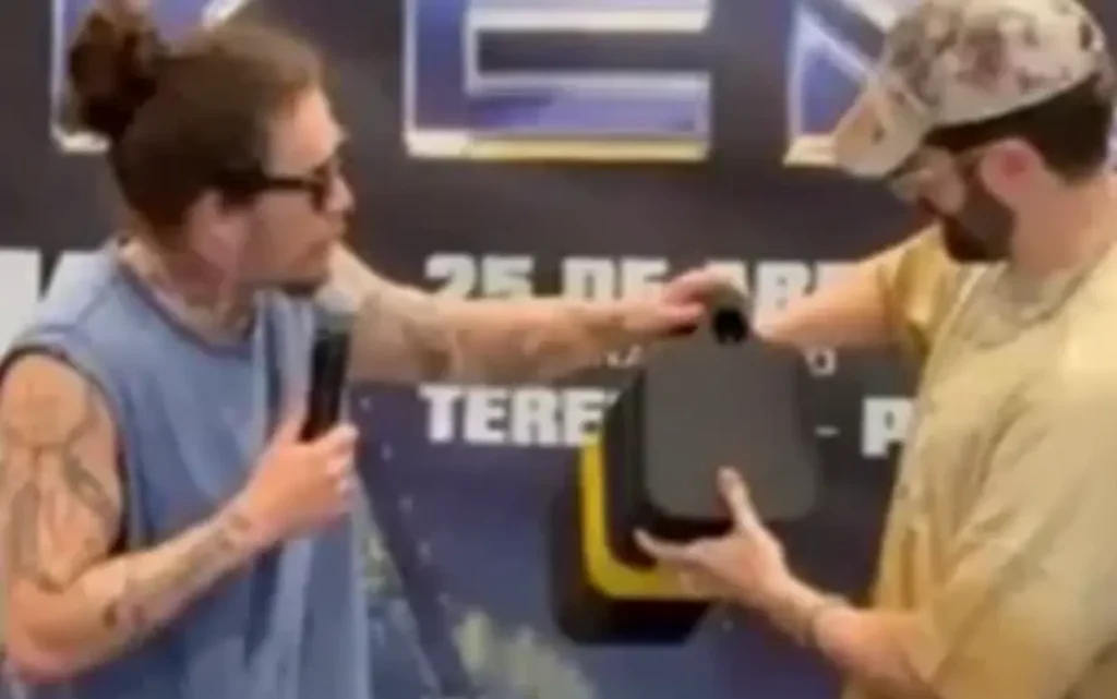 Dois homens em um palco. Um, com cabelo preso e microfone, entrega um objeto preto ao outro, que usa boné camuflado.