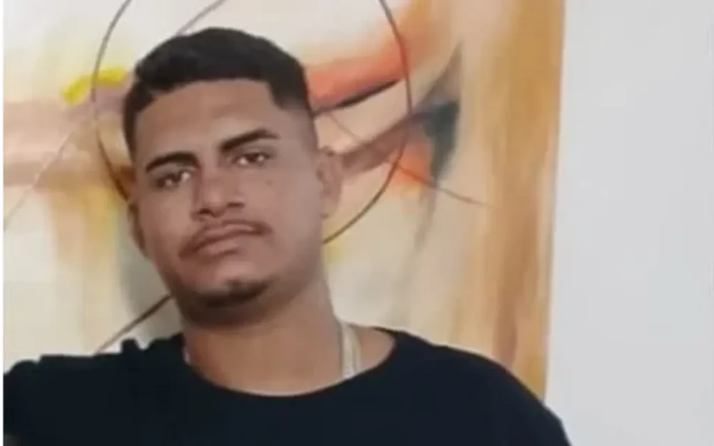 Retrato de um jovem pardo de cabelo curto, bigode e cavanhaque. Ele veste camiseta preta e olha seriamente para a câmera.