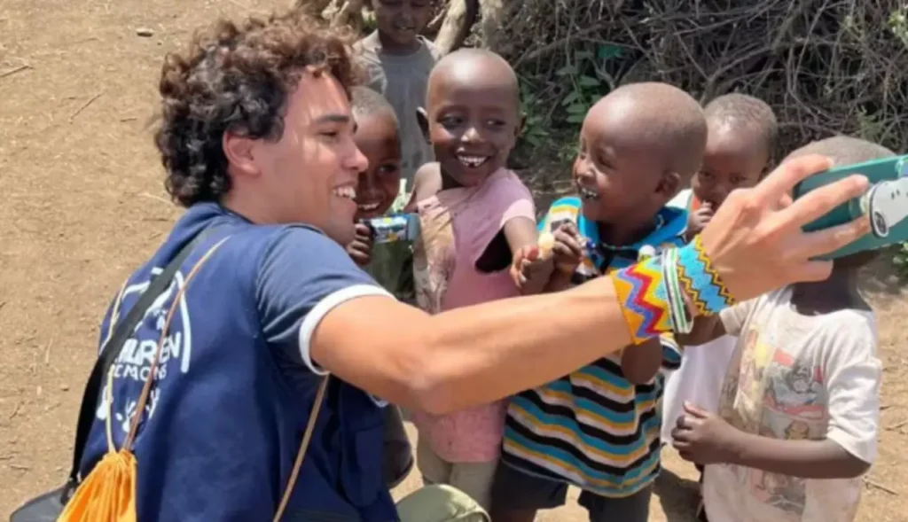 Jovem voluntário de cabelo cacheado tira uma selfie sorridente com um grupo de crianças negras em uma comunidade rural.