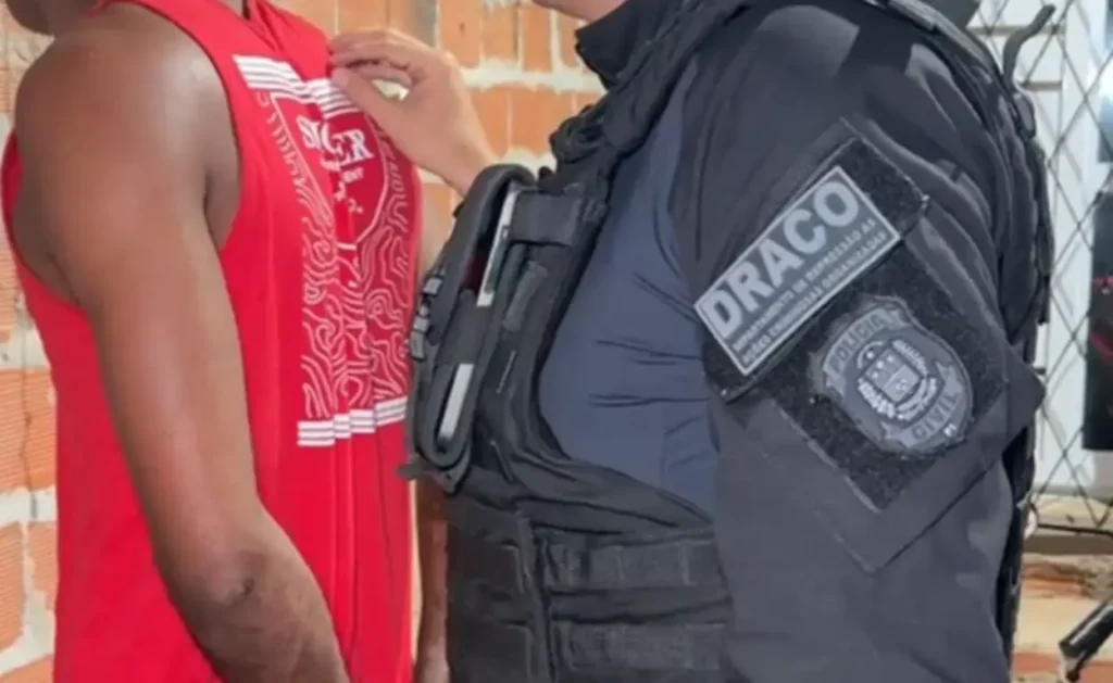 Policial civil do DRACO aborda um homem de camisa regata vermelha, visto de costas, durante uma operação.