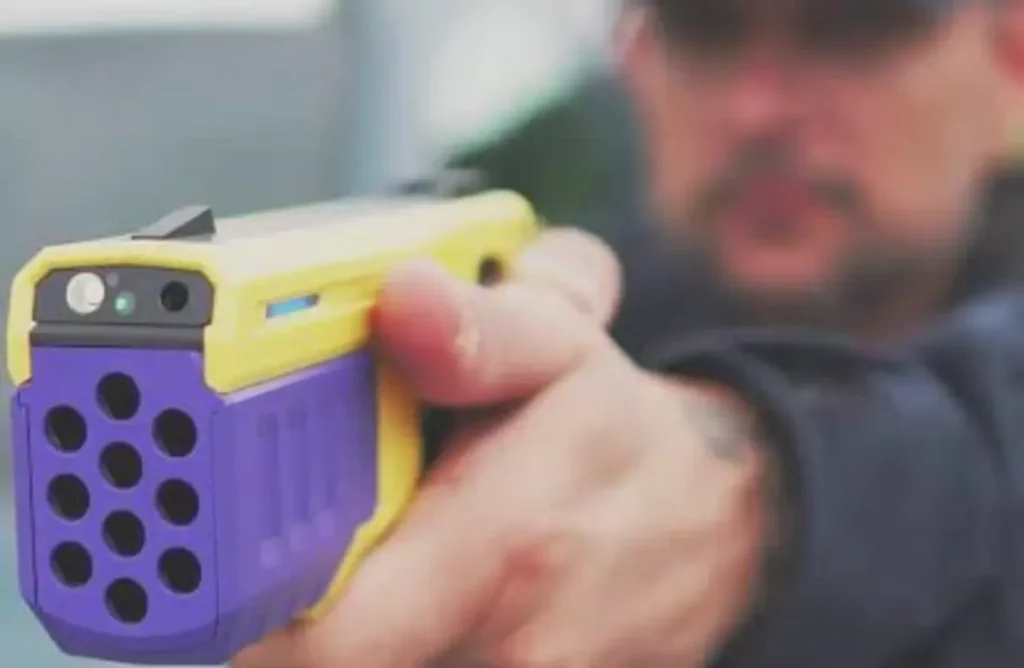 Close-up de uma arma não letal, amarela e roxa, sendo empunhada por um homem com o rosto desfocado ao fundo.