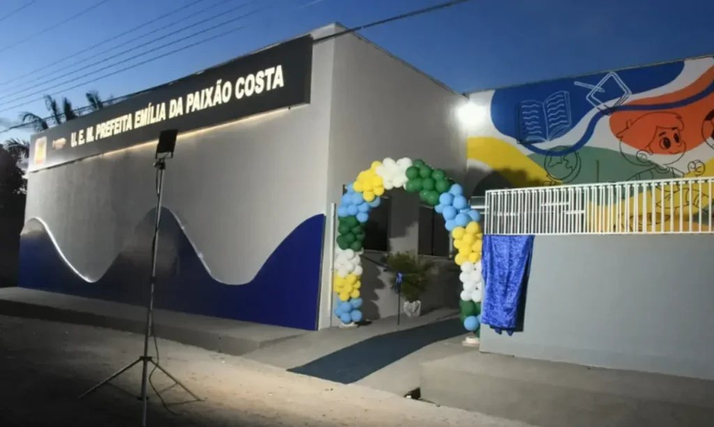 Fachada da escola U.E.M. Prefeita Emília da Paixão Costa à noite, decorada com um arco de balões coloridos na entrada.
