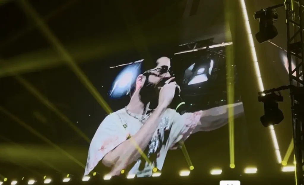 Cantor se apresentando em um telão durante show, com luzes de palco amarelas e estrutura de iluminação visíveis.