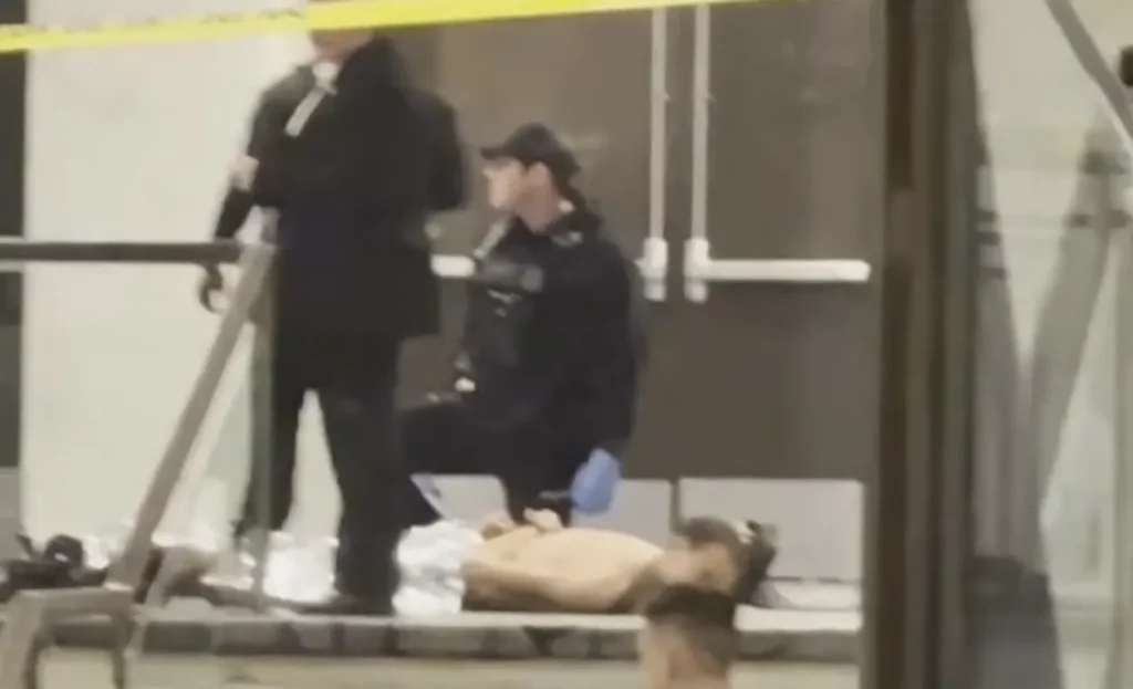 Cena de crime com policial ajoelhado ao lado de uma pessoa deitada no chão, atrás de uma fita de isolamento amarela.