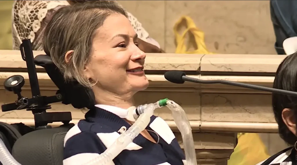 Mulher sorridente em cadeira de rodas com tubo de traqueostomia, falando ao microfone em um ambiente formal.