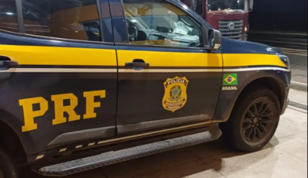 Close-up da lateral de uma viatura da Polícia Rodoviária Federal (PRF), destacando a sigla, o brasão e a bandeira do Brasil.