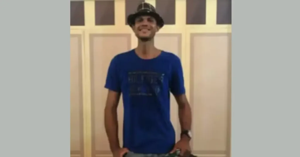 Homem sorrindo para a câmera, usando chapéu xadrez e camiseta azul, em frente a uma parede com painéis claros.