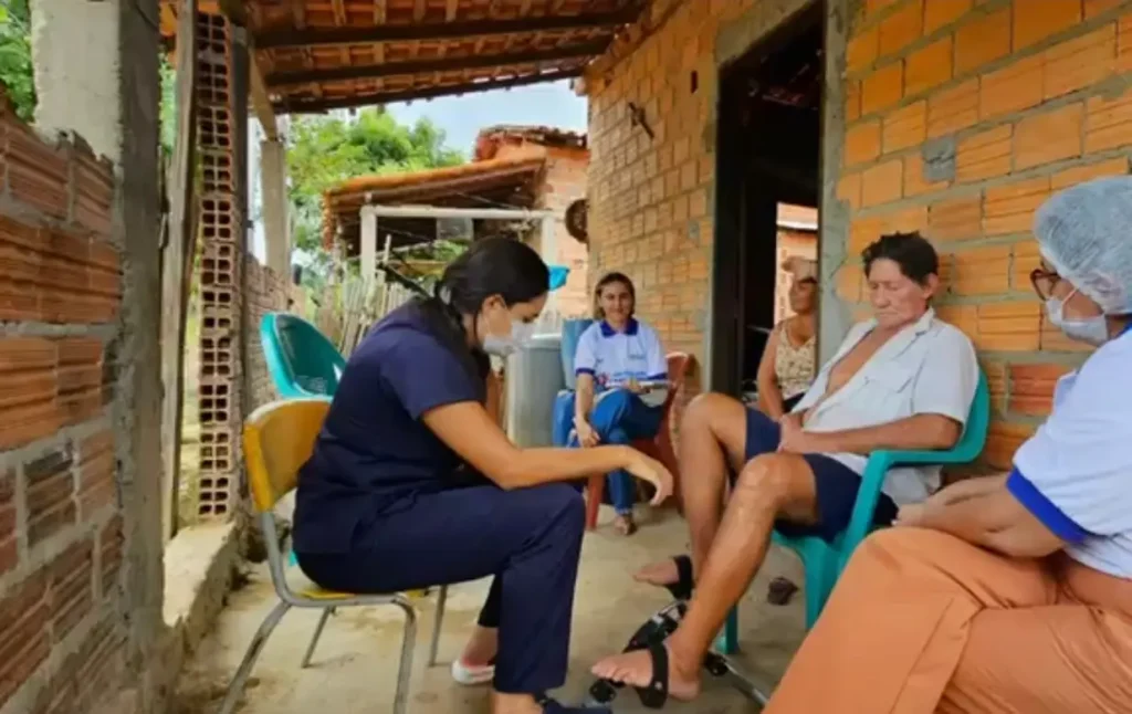 Profissional de saúde de uniforme azul e máscara examina o pé de um homem sentado na varanda de uma casa de tijolos.