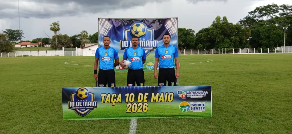 Três árbitros uniformizados com camisa azul e short preto posam em campo gramado segurando uma bola de futebol, à frente de faixa oficial da Taça 10 de Maio 2026, organizada pela Prefeitura de São Francisco do Maranhão e Secretaria de Esporte e Lazer