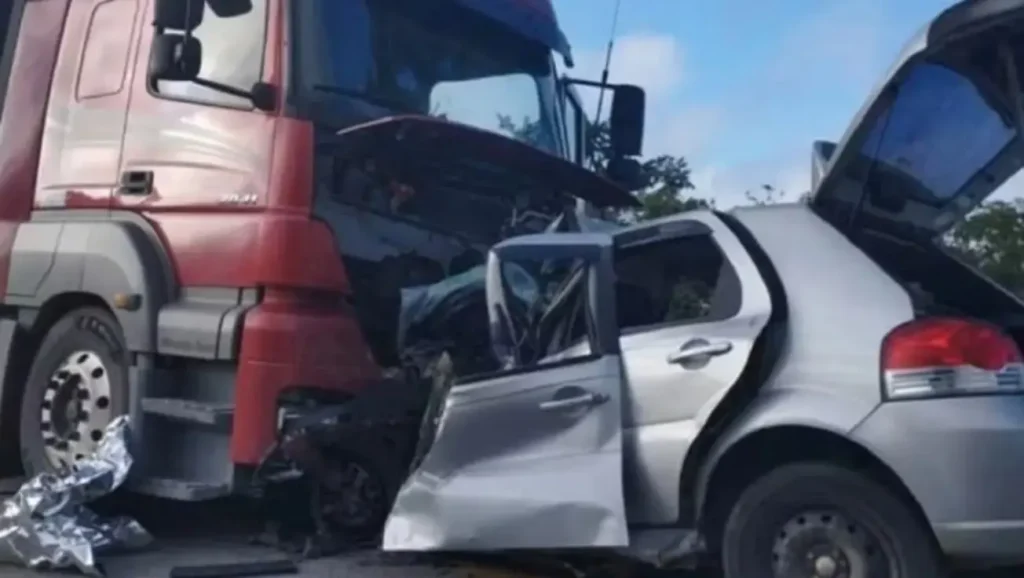 Carro prata com a frente esmagada sob uma carreta vermelha após acidente. A porta do motorista está aberta e amassada.