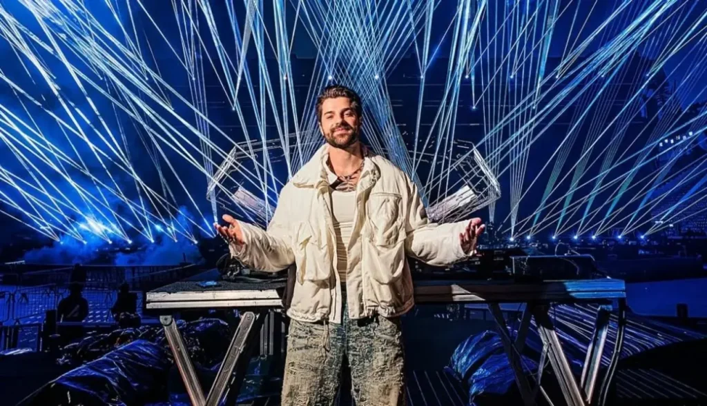 DJ Alok sorrindo de braços abertos no palco, em frente a equipamentos de som, com um show de lasers azuis ao fundo.