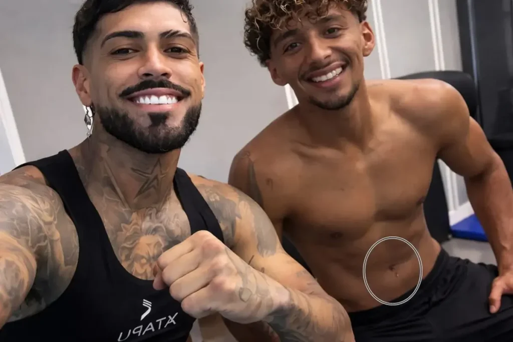 Dois homens jovens e musculosos posam para selfie em academia: o da esquerda veste regata preta com tatuagens no corpo e braços, fazendo gesto de soco; o da direita está sem camisa, exibindo abdômen definido com um círculo branco destacando a região abdominal