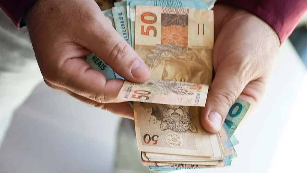 Aposentado contando cédulas segurando cédulas do pagamento do 13º salário antecipado 2026