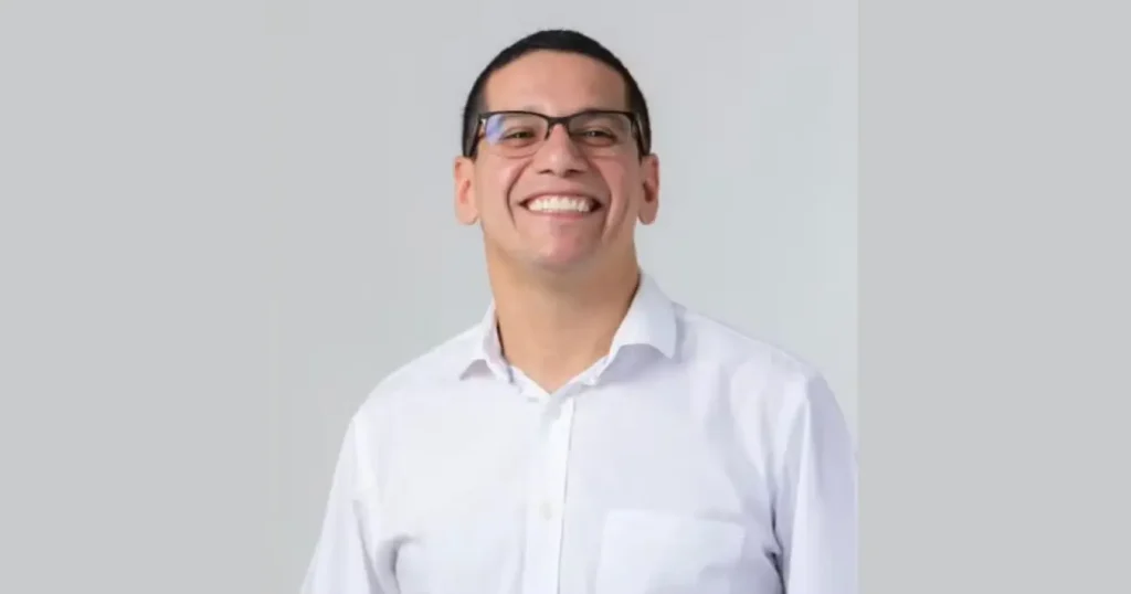 Foto de Daniel Oliveira, um homem de óculos e camisa branca, sorrindo para a câmera com um fundo cinza claro.