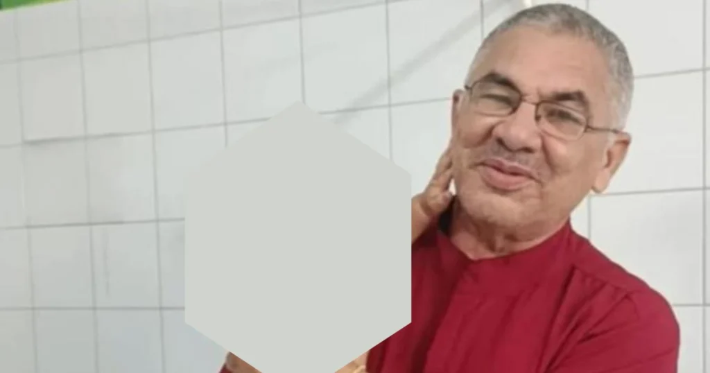 Homem de óculos e camisa vermelha, com cabelo grisalho, posa com a mão no pescoço em frente a uma parede de azulejos.