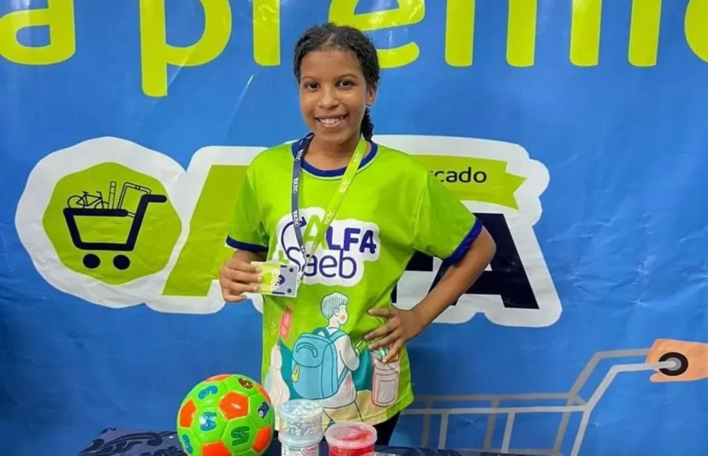 Uma professora sorridente auxilia um menino, aluno da rede pública, a escrever em um caderno em uma sala de aula clara e bem iluminada.