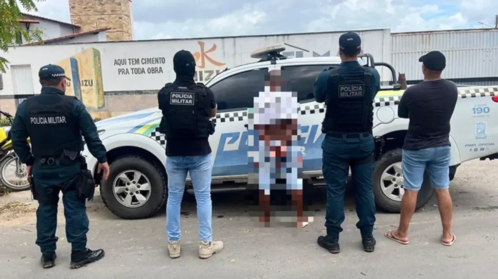 Policiais militares ao lado de viatura durante a detenção de um suspeito, que está sentado no chão com a imagem pixelada.