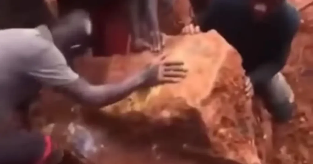 Garimpeiros se reúnem ao redor de uma enorme pedra dourada, que aparenta ser uma pepita de ouro, em um buraco de terra.