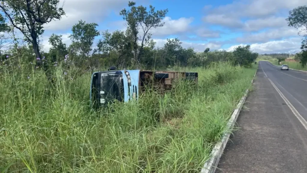 Ônibus tombado na lateral de uma rodovia, em meio à vegetação alta, com um carro ao fundo e céu azul com nuvens.