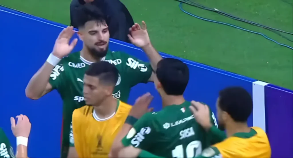 Jogadores do Palmeiras com uniformes verde e branco se abraçam em comemoração de gol, com um jogador de uniforme amarelo ao centro da cena, próximos à placa azul de beira de campo