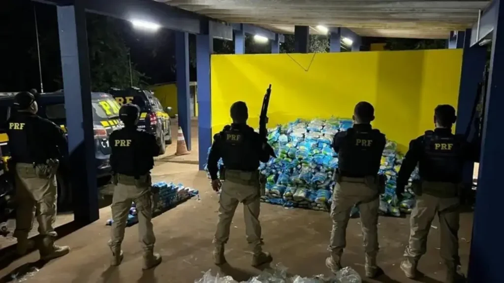 Agentes da PRF, um deles segurando um fuzil, fazem a guarda de uma grande pilha de pacotes de droga apreendida à noite.