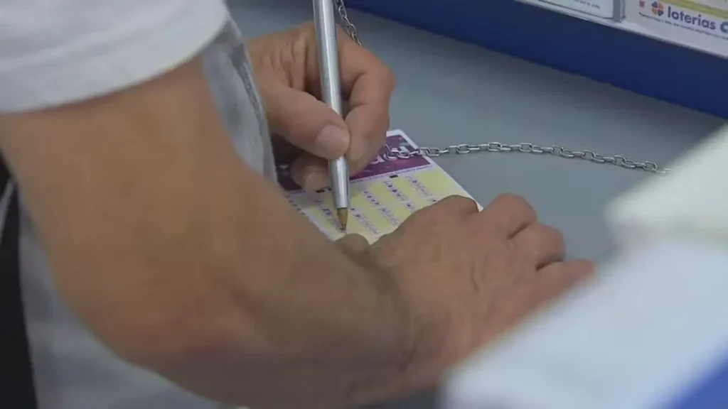 Mãos de uma pessoa segurando uma caneta e preenchendo um volante de aposta da Lotofácil sobre um balcão de lotérica.