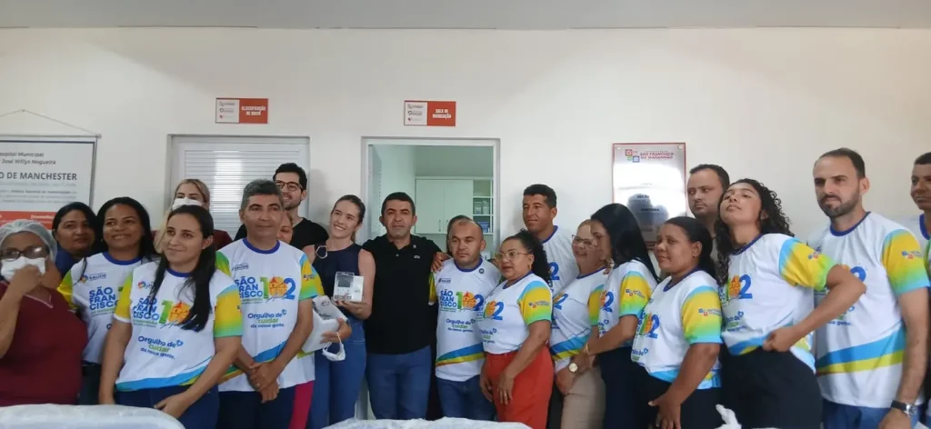 Grupo de pessoas em t-shirts brancas e coloridas posando em ambiente interno, uma mulher segura aparelho médico.