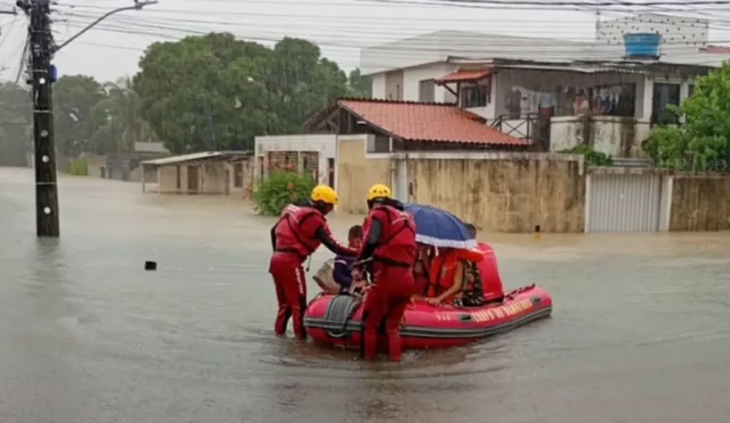 Resgatistas em coletes vermelhos e capacetes amarelos ajudam pessoas, incluindo crianças, em bote inflável vermelho em rua alagada por causa das chuvas em Pernambuco