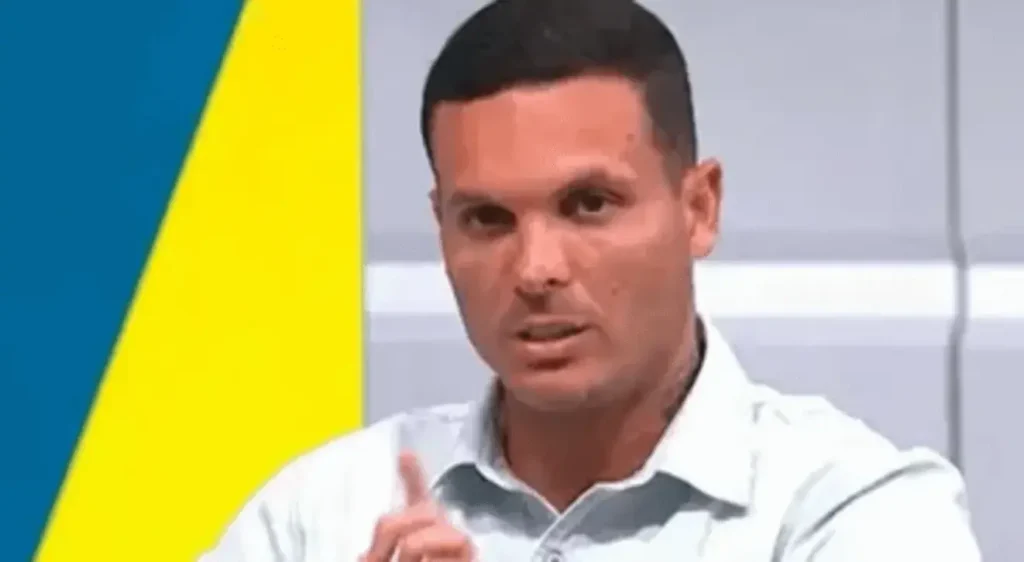 Homem de camisa clara, cabelo escuro, apontando o dedo indicador para cima, em fundo azul, amarelo e cinza de estúdio.