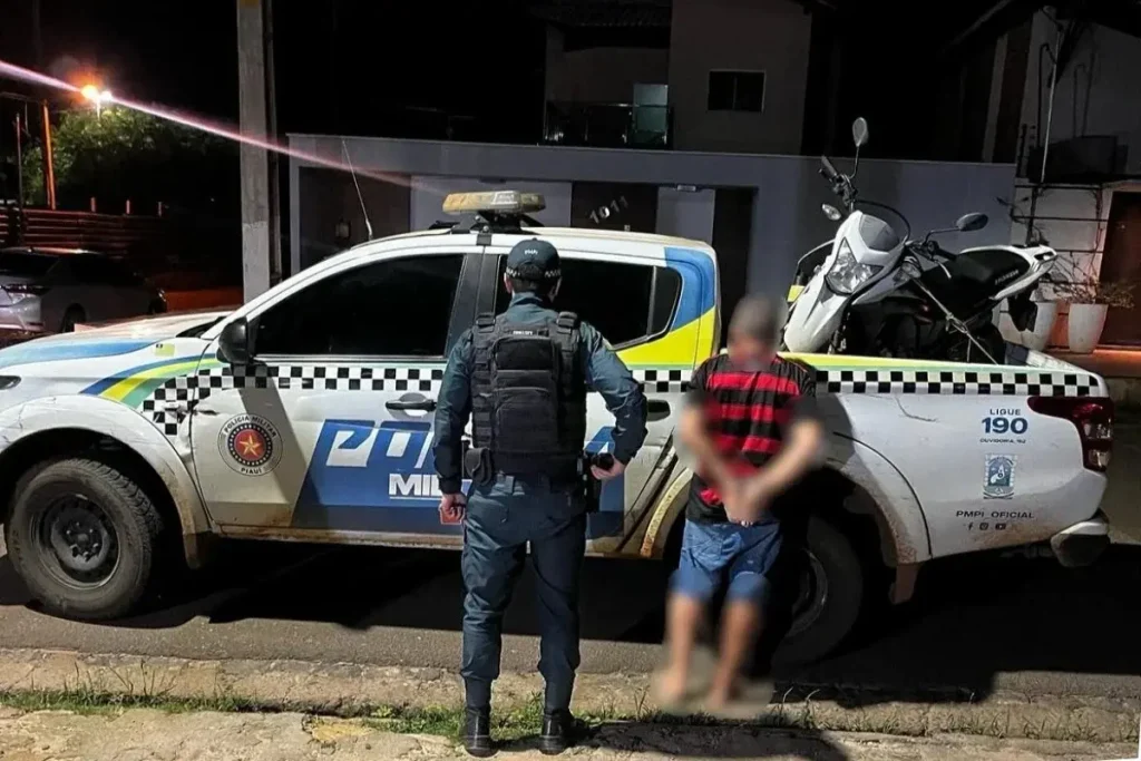 Policial militar e motociclista preso em barreira com imagem borrada ao lado de viatura com moto apreendida na caçamba, à noite.