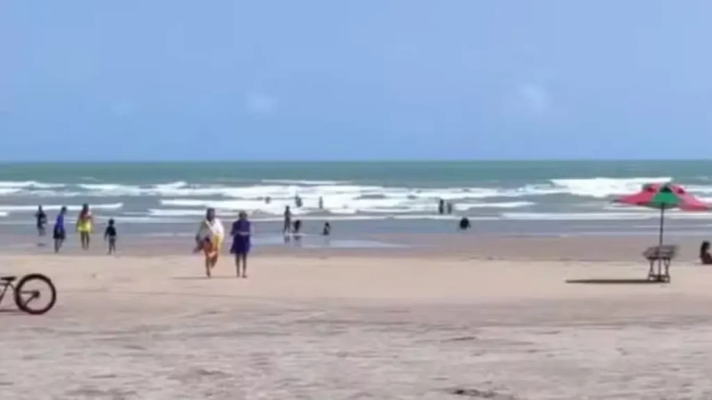 Pessoas na praia com mar agitado, céu azul, guarda-sol e bicicleta. Cena de lazer em dia ensolarado.