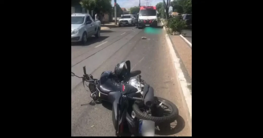 Motocicleta caída no asfalto após acidente, com capacete e ambulância ao fundo em via urbana.
