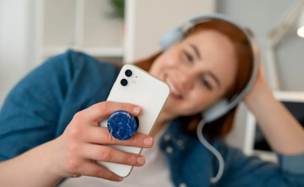 Jovem com fones de ouvido segurando smartphone branco com suporte pop socket azul marmorizado fixado na parte traseira
