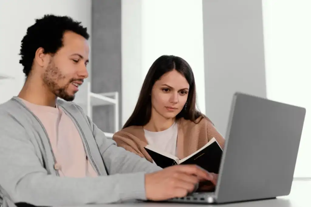 Duas pessoas colaborando: um homem digita no notebook enquanto segura um caderno, e uma mulher observa a tela atentamente