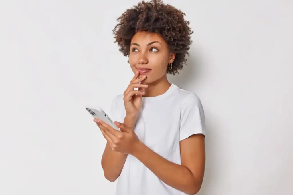 Mulher jovem com cabelo cacheado segura um smartphone e olha pensativa para o lado, com o dedo no queixo