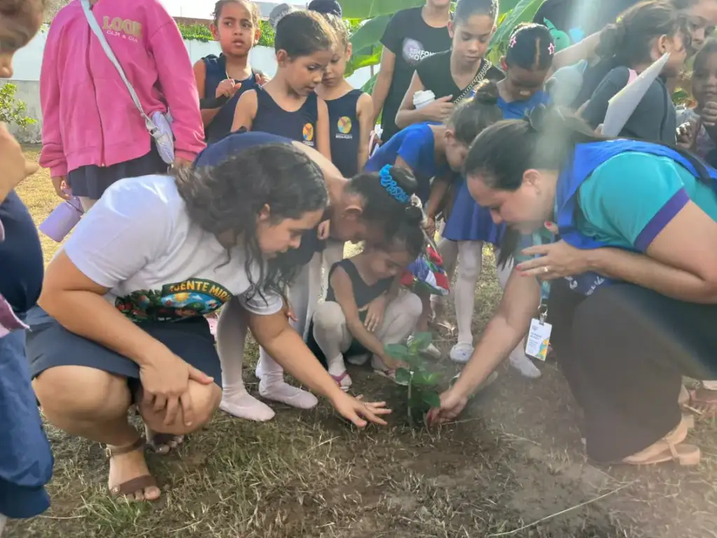 Crianças e adultos agachados plantando uma muda no solo, em atividade educativa ao ar livre