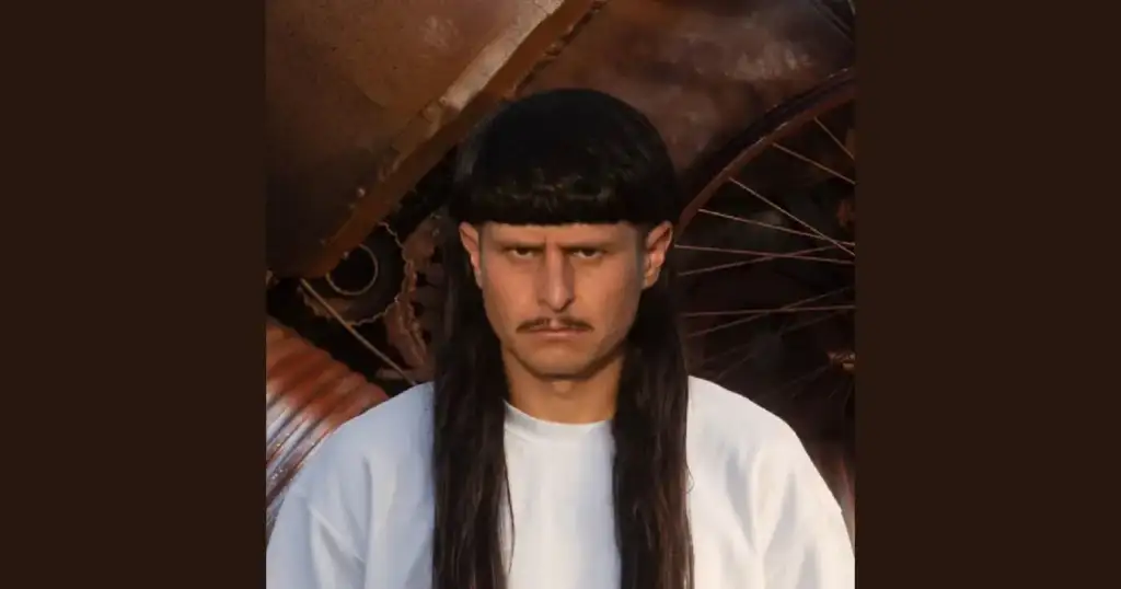 Cantor americano Oliver Tree com cabelo preto longo e franja reta, bigode fino e expressão séria, vestindo camiseta branca, com rodas enferrujadas ao fundo