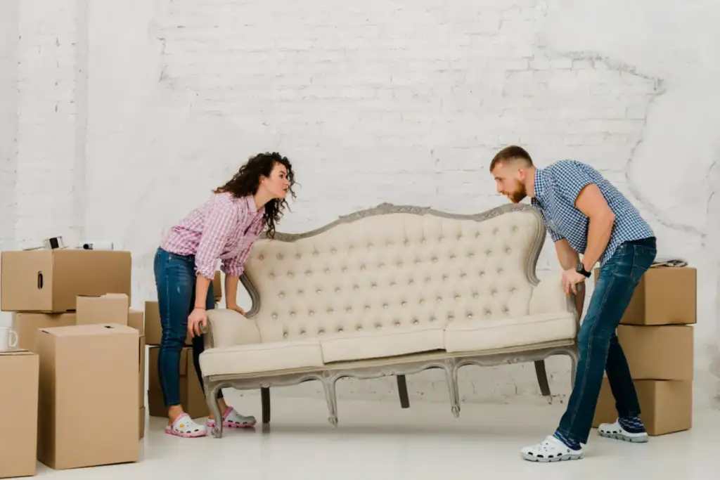 Casal carregando um sofá clássico bege em meio a caixas de papelão durante uma mudança