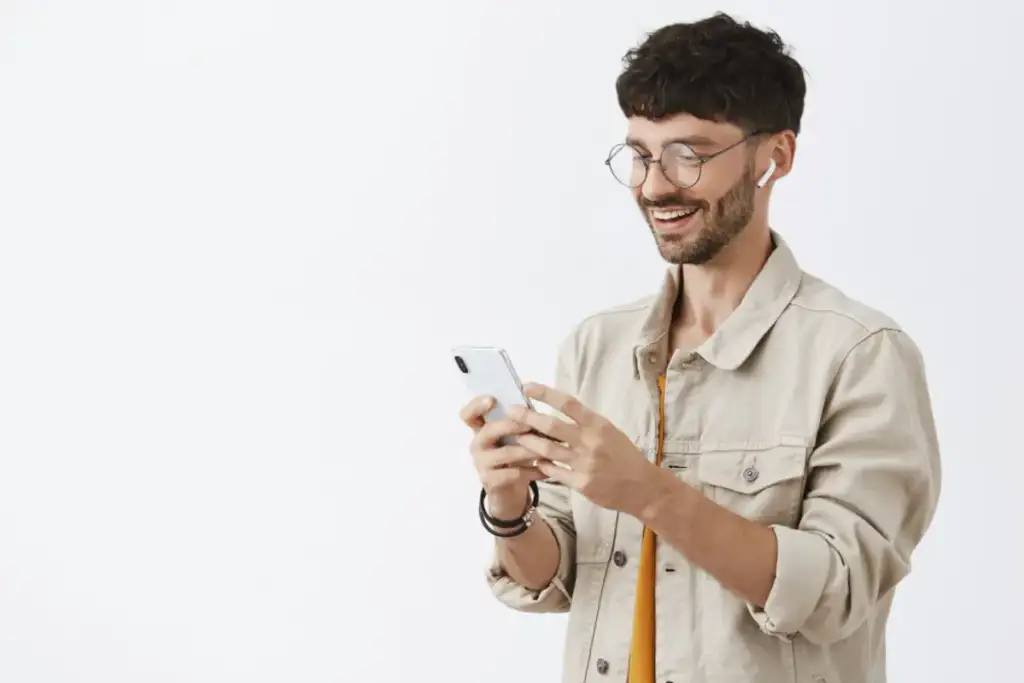 Homem jovem sorrindo enquanto usa o smartphone, com fone de ouvido sem fio e óculos redondos