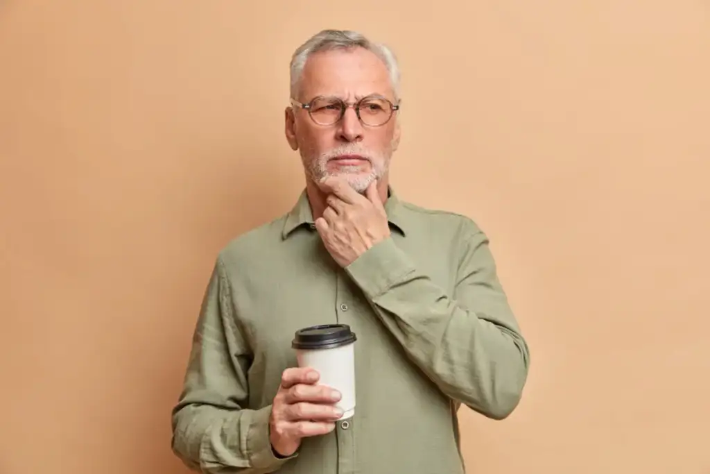 Homem idoso de óculos e cabelos grisalhos segura copo de café descartável com expressão pensativa