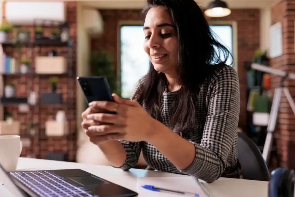 Mulher sorrindo enquanto usa o smartphone em uma mesa de trabalho com notebook e caderno ao fundo