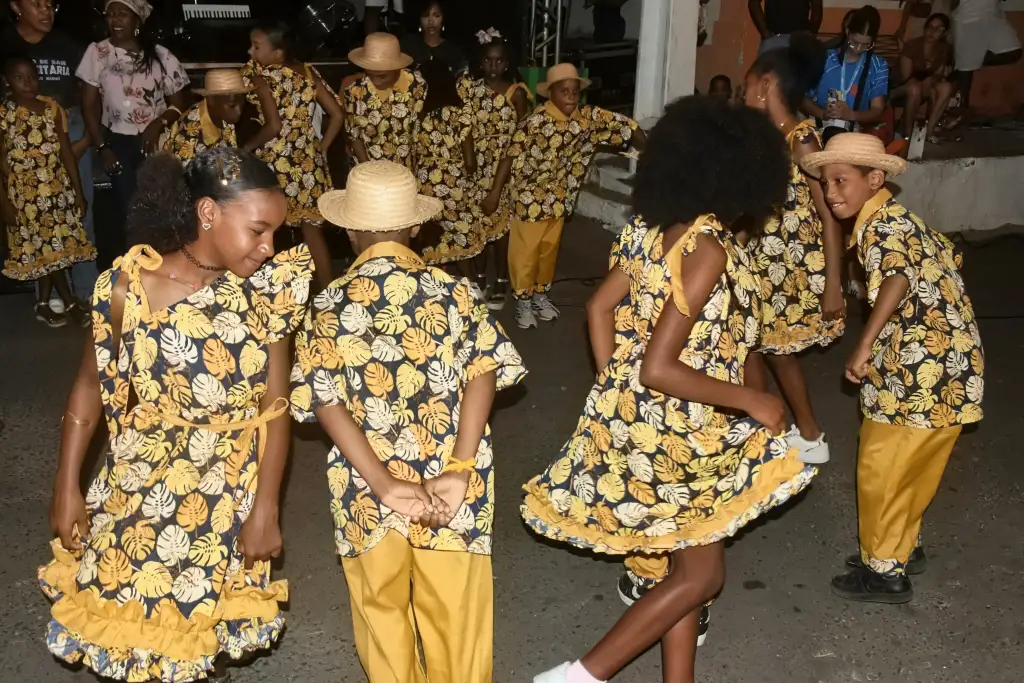 Crianças com trajes típicos amarelos e azuis dançando em apresentação de festa junina ao ar livre à noite