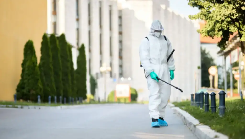 Profissional com macacão de proteção branco, máscara e luvas realiza desinfecção em área urbana com equipamento de pulverização